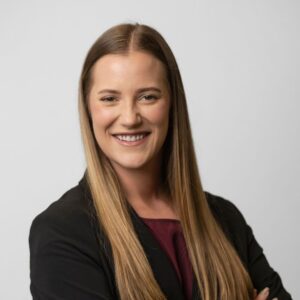 A woman with long straight hair, wearing a black blazer over a burgundy top, smiles confidently with arms crossed against a plain light background.