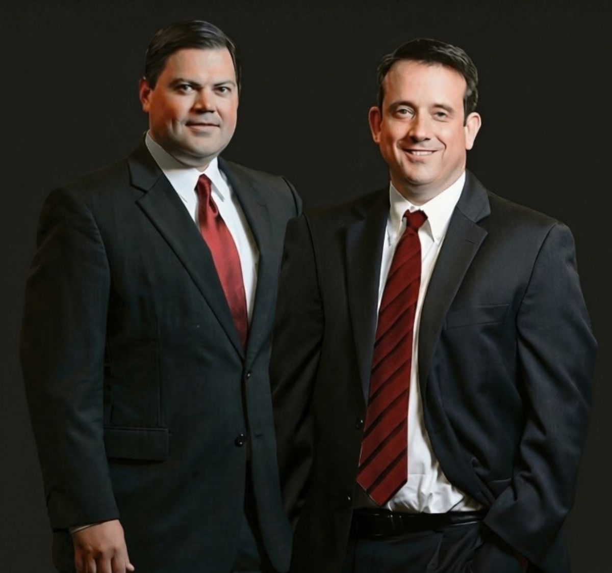 Two men in dark suits and red ties stand side by side against a plain black background, both facing forward and smiling slightly.
